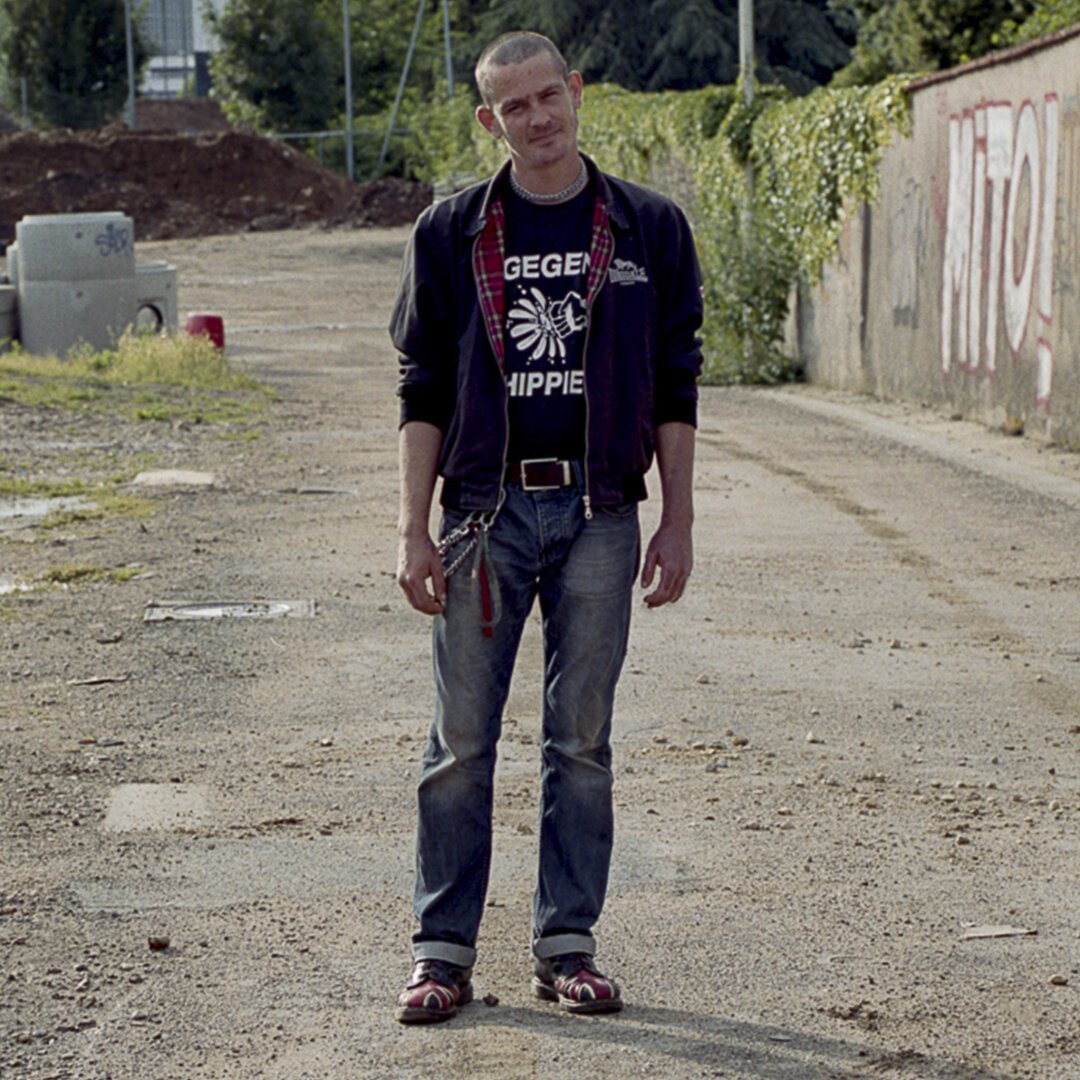 Franck, un homme d'une trentaine d'année, est débout, plutôt souriant, il porte un veste Lonsdale, un jean et des Dr Martens. En arrière plan un terrain vague