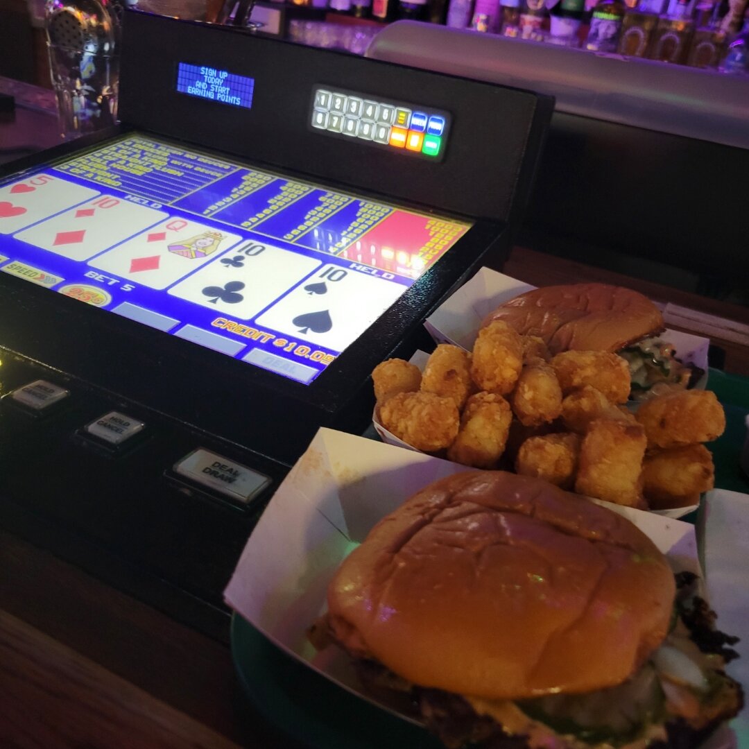 A  video poker machine displaying cards on screen sits next to a plate of food containing a large burger and golden fried tots or nuggets. The gaming machine shows playing cards including a 6 of clubs and 10 of spades, with colorful bar atmosphere lighting in the background.