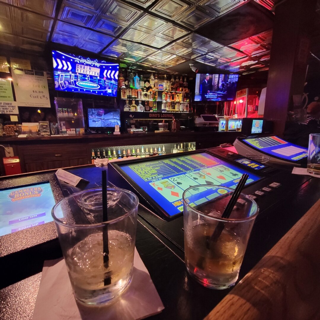 Interior of a dimly lit bar with decorative tin ceiling tiles and colorful LED lighting. In the foreground are two cocktails with black straws on a dark table, next to video poker machines displaying card games. The bar area features multiple TV screens, shelves of liquor bottles, and gaming machines, all illuminated by purple, blue, and red ambient lighting.