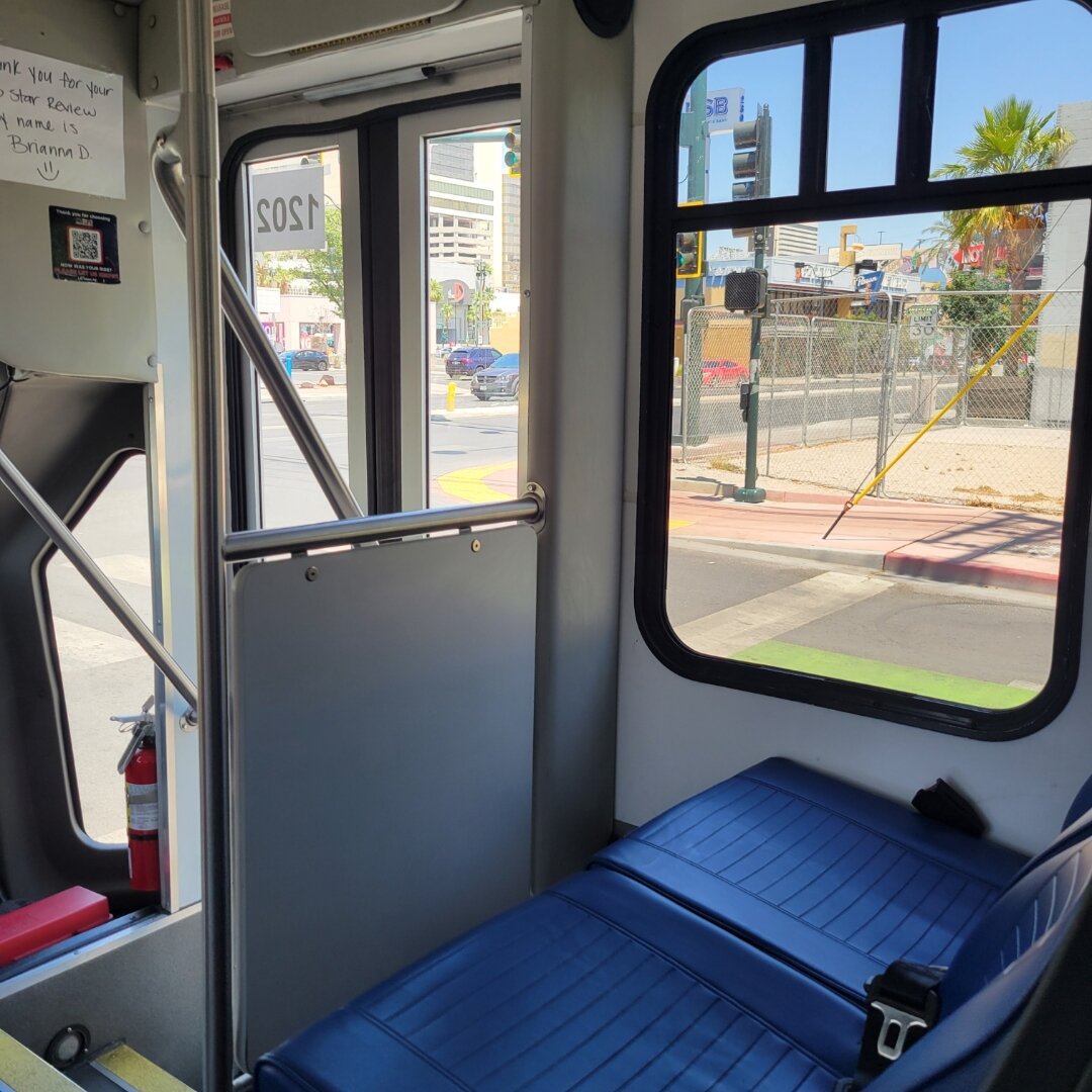 Interior view of a public bus showing blue vinyl seats, metal handrails, and large windows. Through the windows, you can see an urban street scene with palm trees, buildings, parked cars, and a chain-link fence. There's a handwritten note visible on the wall inside the bus