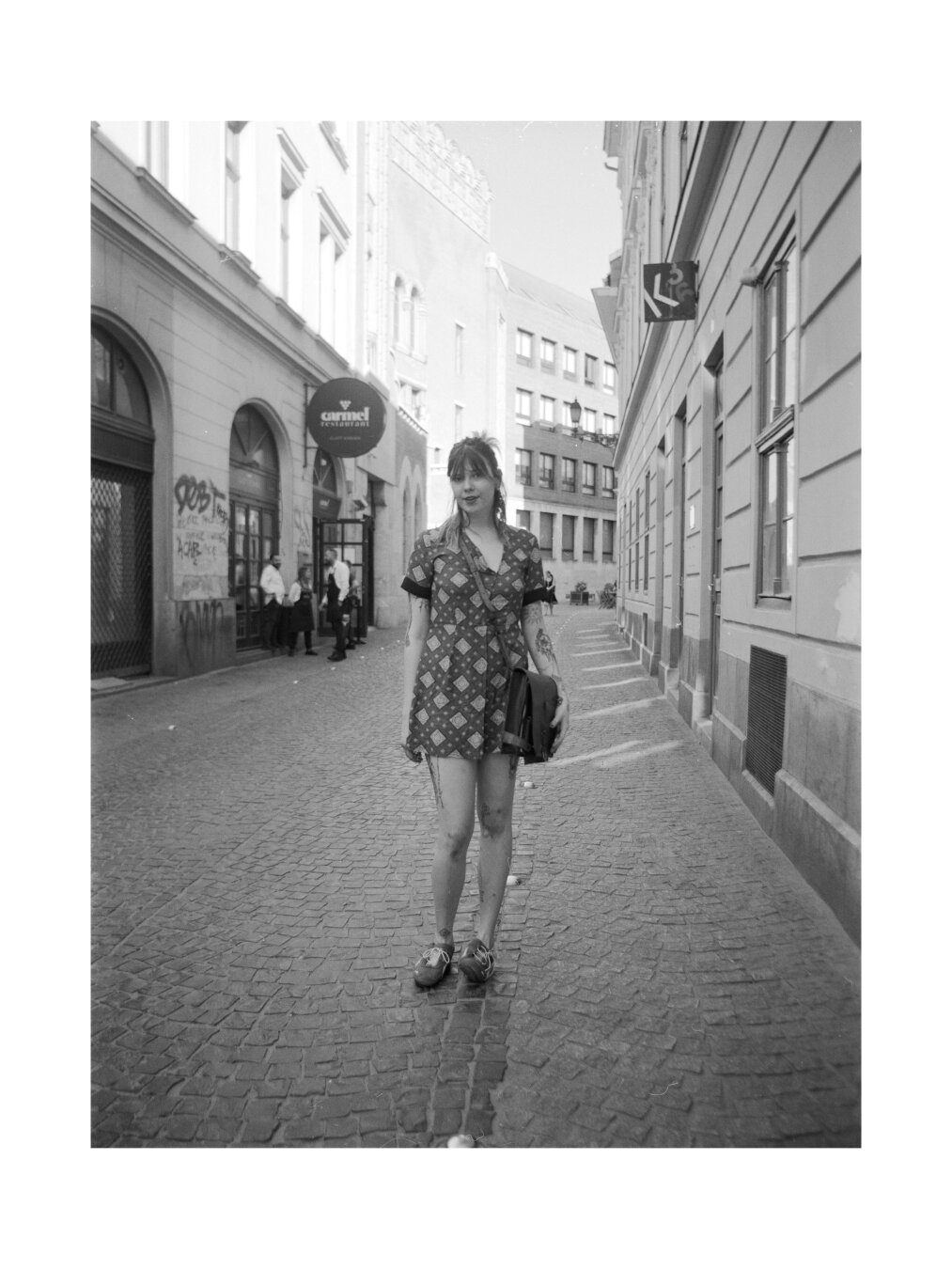 A black and white photo of a women in short dress. The background is a historical street of a big city. In the left of the image in the background there are a group of waiters.