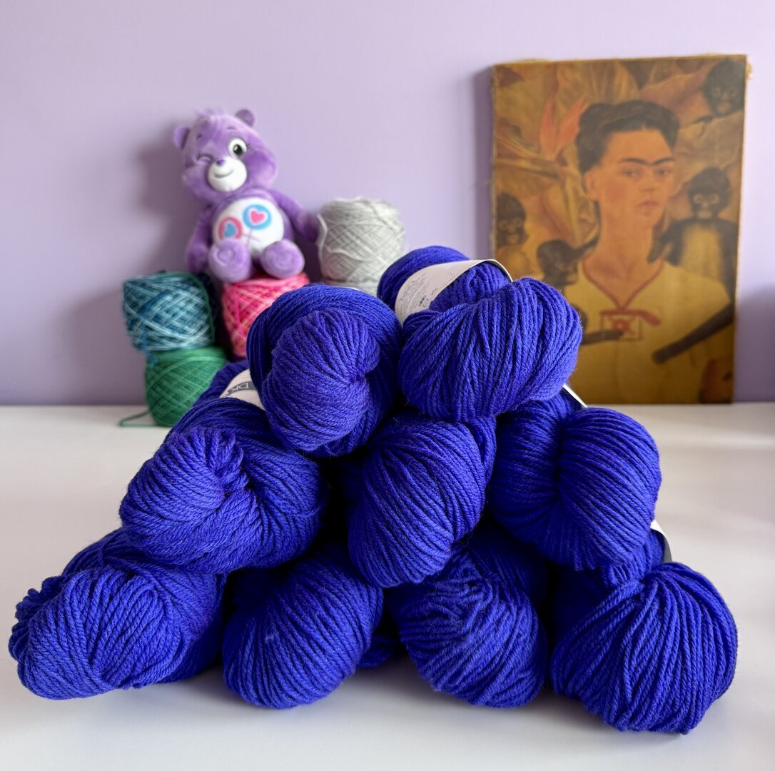 A stack of 9 skeins of yarn in a dark neon almost blue purple color, in the background are balls of yarn with a care bear on it and a canvas with Frida kahlo against a purple wall