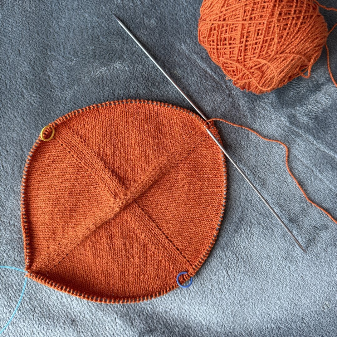 Top down view of an orange center-out circular knit fabric with increases at 4  different points. A ball of orange yarn sits next to the work in progress.