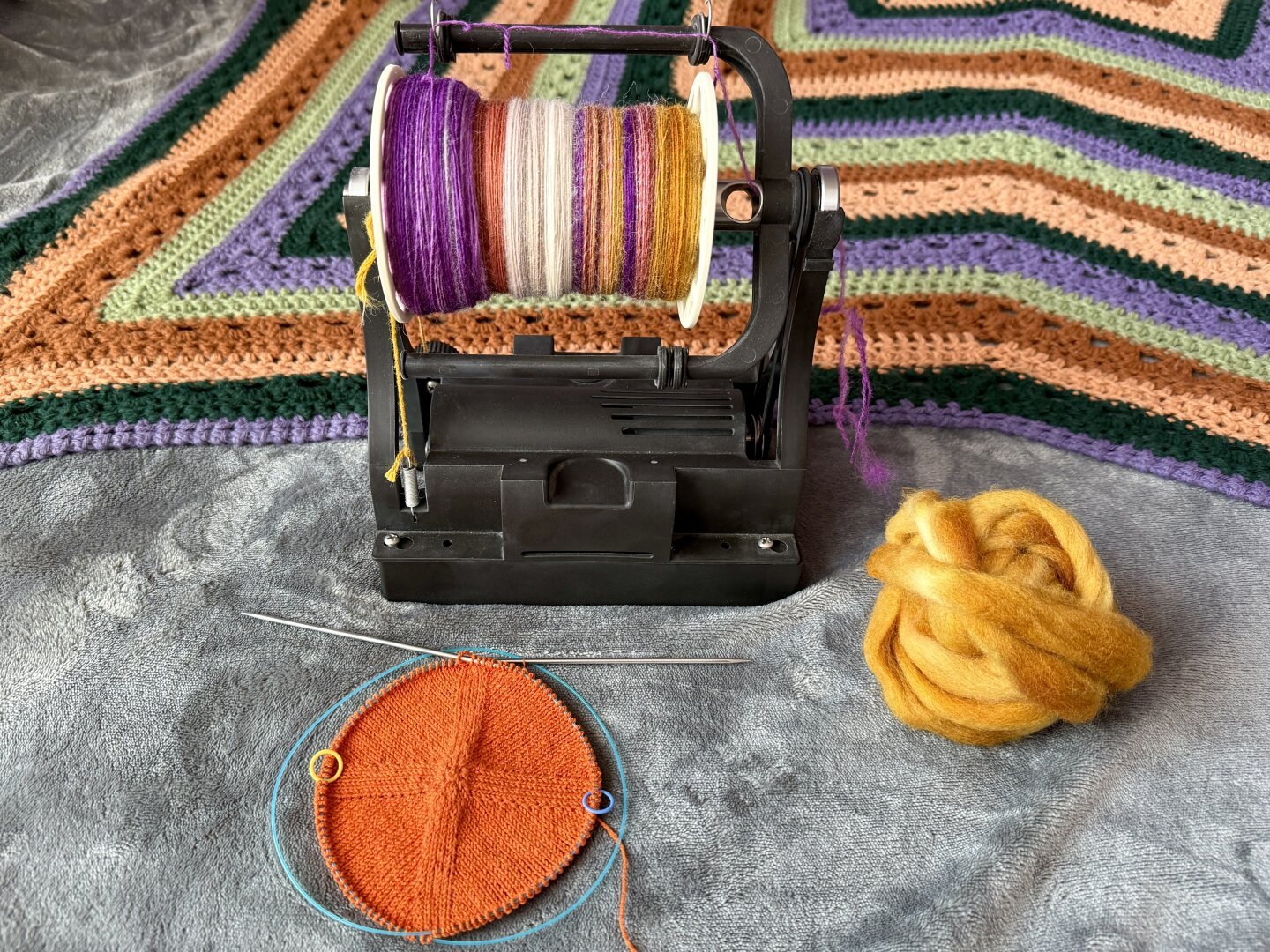 An orange knit hat in progress laying flat to show its circular shape. It sits in front on an electric spinning wheel with yarn in progress in the bobbin in a combination of purple, grey, yellow, copper colors. Next to the spindle is a bundle of yellow fiber for spinning. In the background is a crocheted striped blanket in purple, green, beige, and brown