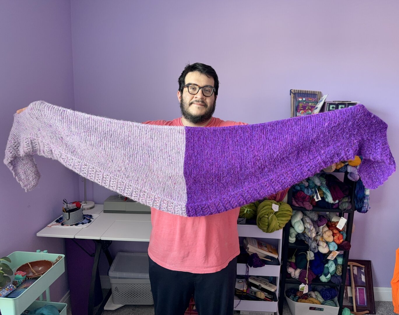 Me holding a crescent shaped shawl with a lace pattern along the bottom border. The left half is grey and pink with tones of pastel purple. The right half is purple with neon purple accents. The shawl has a fuzzy texture.