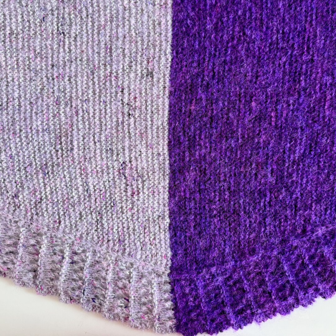 The center of a crescent shaped shawl showing two halves in different colors. On the left is a grey and pink work pink boucle, on the right is purple with neon purple boucle.