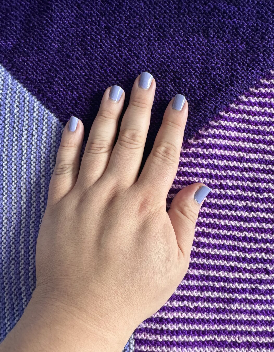 My hand placed on a modular dark purple, periwinkle, and magenta shawl. My nails have a lilac/lavender polish on them