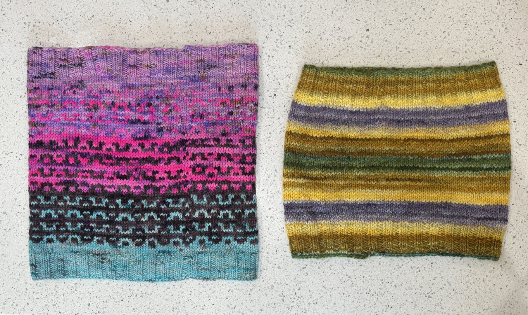 Two cowls laying flat. The left cowl has light blue, hot pink and purple sections with mosaic colorwork in black pink and purple. The right cowl has stripes in purple, yellow, brown, and green that comes from the transitions in the yarn itself.