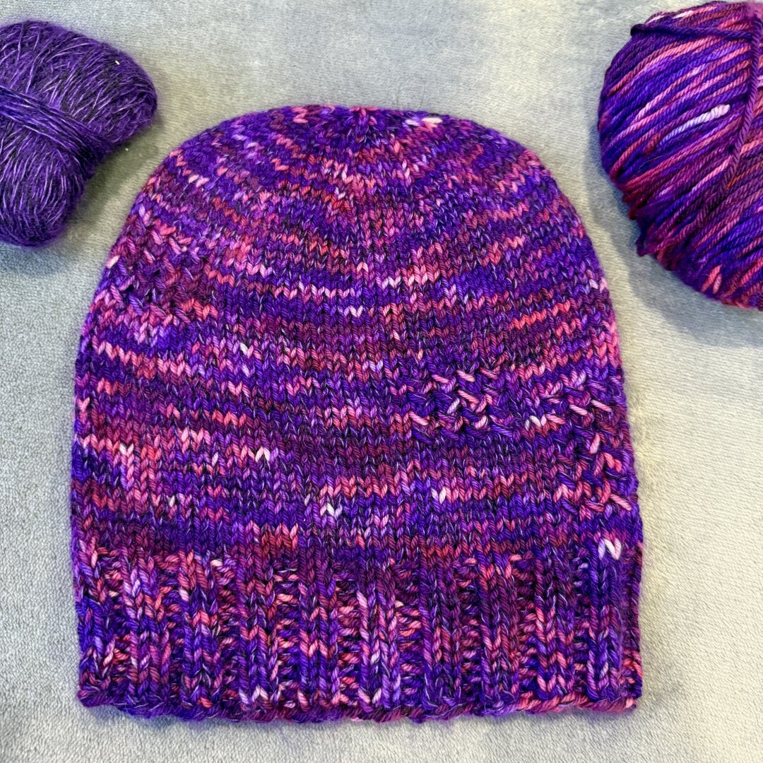 A finished knit hat laying on a blanket. The color is purple with pink, white and blue variegations and it has flecks of a deep purple. To the left of the hat is a thinner deep purple yarn with a slight fuzz, to the right is a thicker yarn that is purple with the pink, white, and blue variegations.