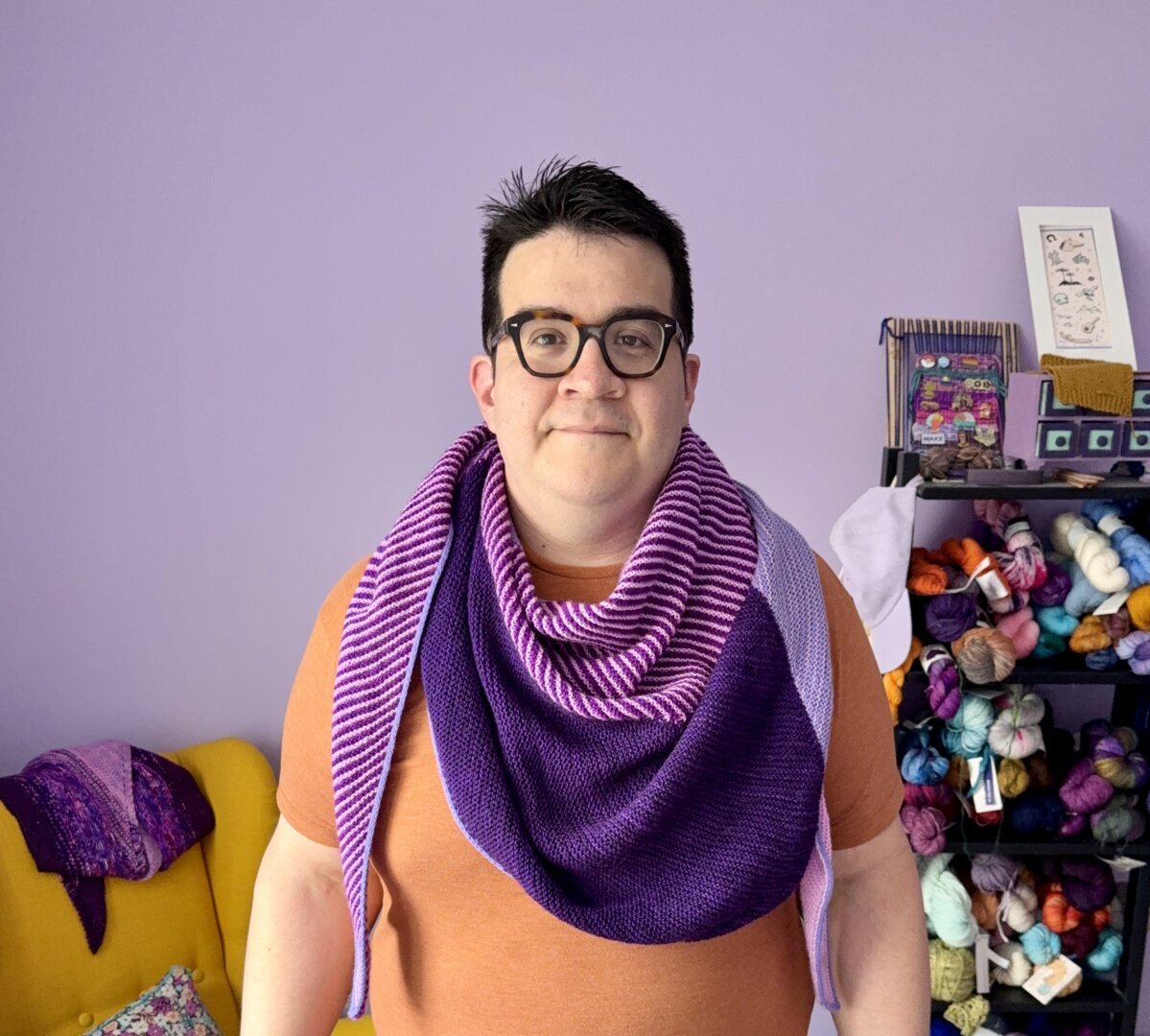 Me wearing modular triangular shawl around my neck, it is made up of grape purple, periwinkle, and striped magenta and lavender sections. I am standing in front of a purple wall, a shelf with yarns and a yellow chair with a purple shawl on it are visible.