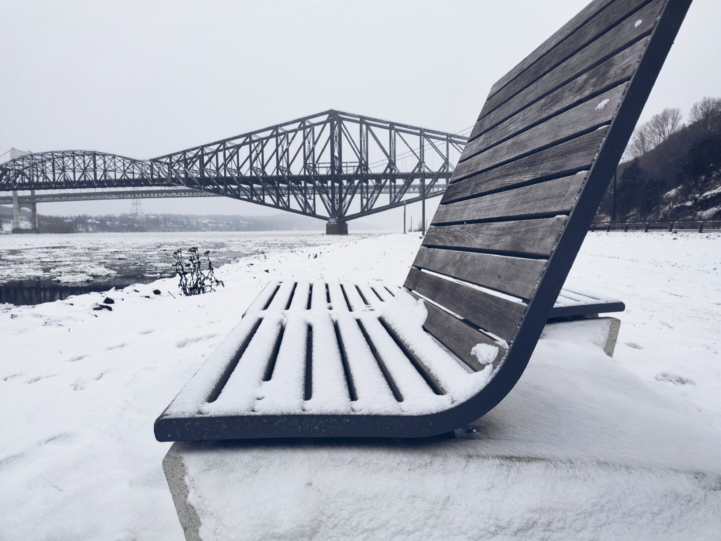 Uno de los varios bancos públicos de madera al borde del río a lo largo de la Promenade Champlain. Al fondo, el puente de Québec en remache de acero.