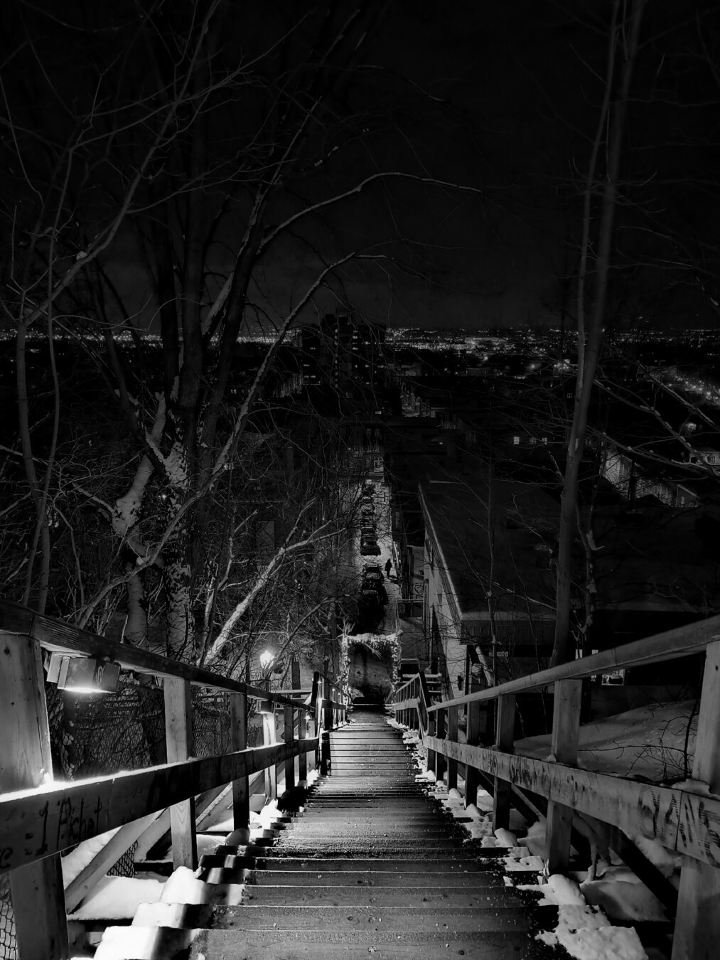 Una de las múltiples escaleras, en este caso de madera, que comunican la parte alta y baja de la ciudad. A los costados hay parches de nieve. Al fondo, una callecita nevada con carros estacionados y una persona caminando. Aún más lejos, las luces nocturnas de la ciudad. Imagen en b/n.