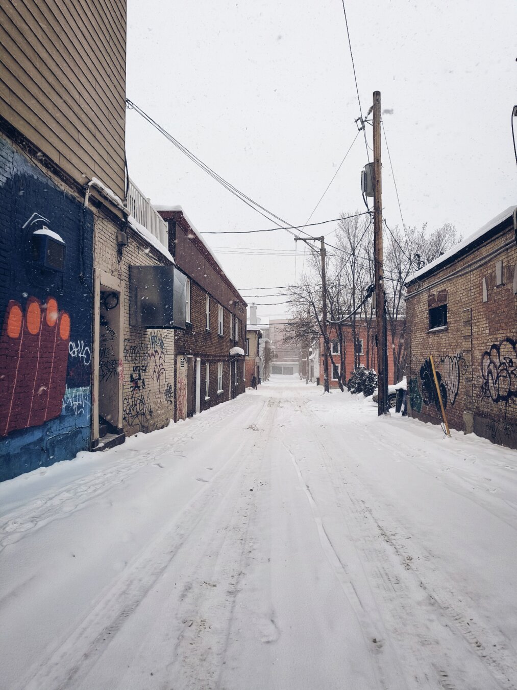 Vista de una calle tranquila, nevada, con trazos de llantas de automóvil enmedio, nevando a media tarde, flanqueada de  edificios viejos de ladrillo, de no más de dos pisos, uno que otro grafiteado y cables y postes de luz. De lejos, una persona camina acercándose, dispuesta a dar vuelta a la derecha.