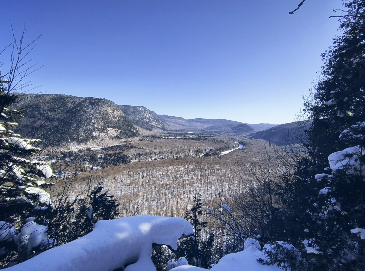 Vista de la Vallée du Bras-du-Nord atravesada por un riachuelo congelado rodeado de montañas viejas cubiertas de árboles deshojados y algo de nieve.