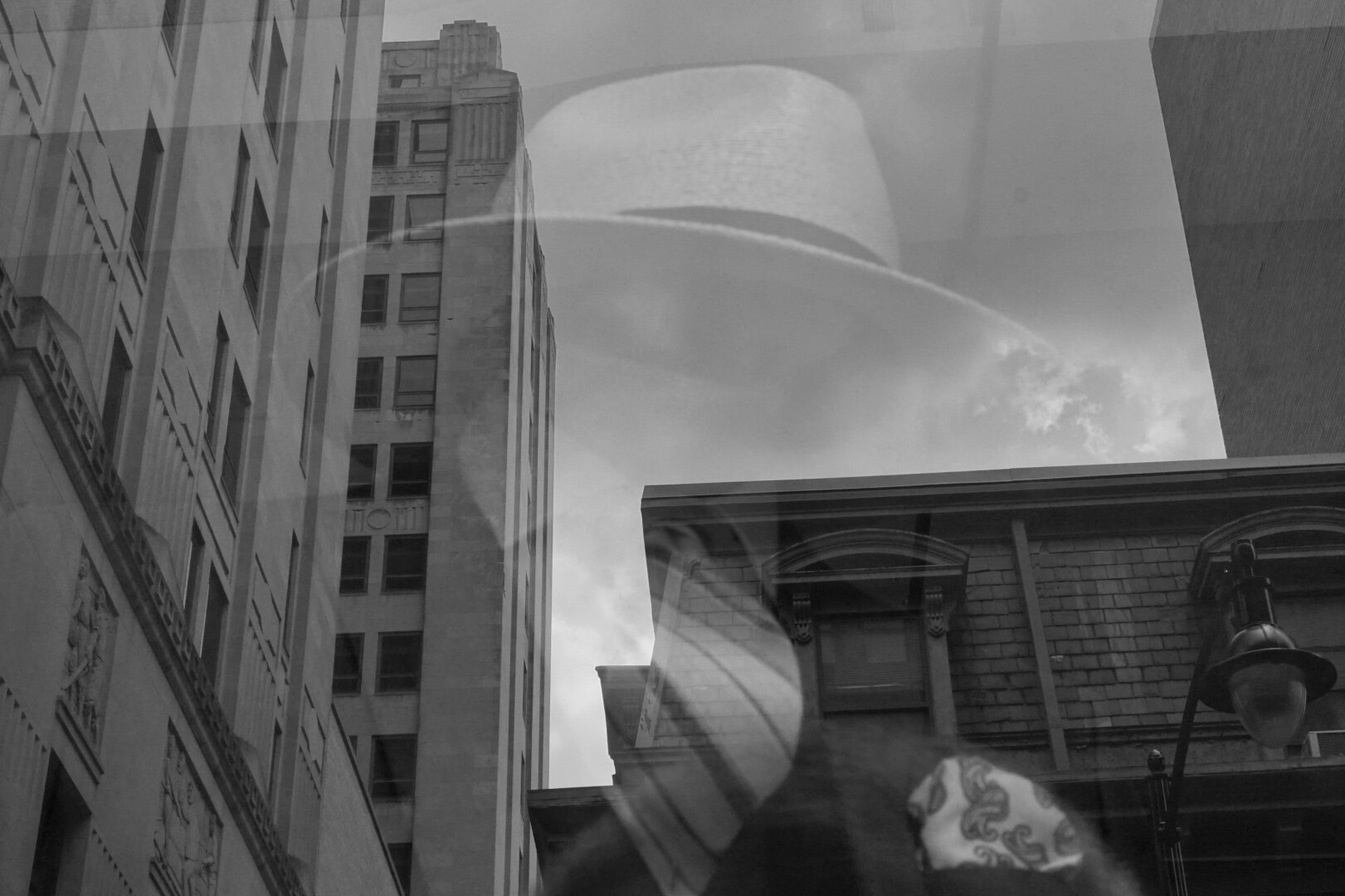 Black and white photograph of a shop window. City buildings are reflected in the window, and blended into the reflection is a mannequin with a fedora and a striped tie. No face is visible.