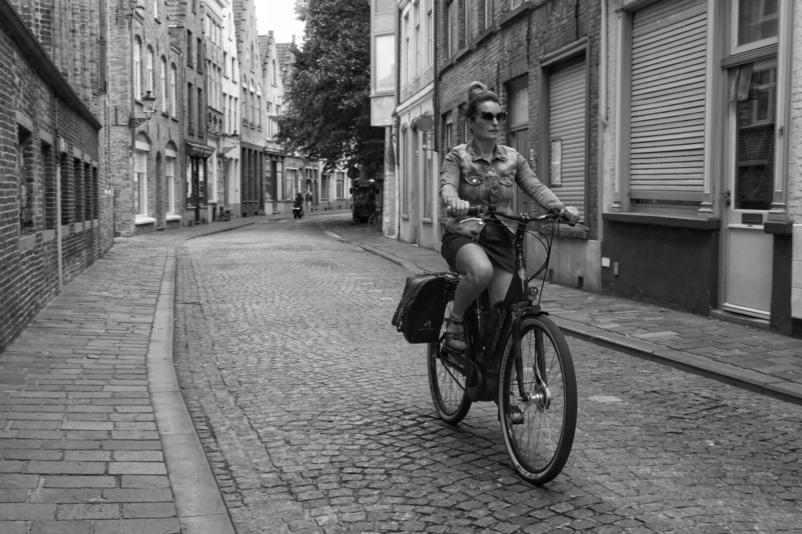 Black and white photo of a woman riding a bicycle down a cobblestone street lined with Old World buildings. The street bends away in the distance.