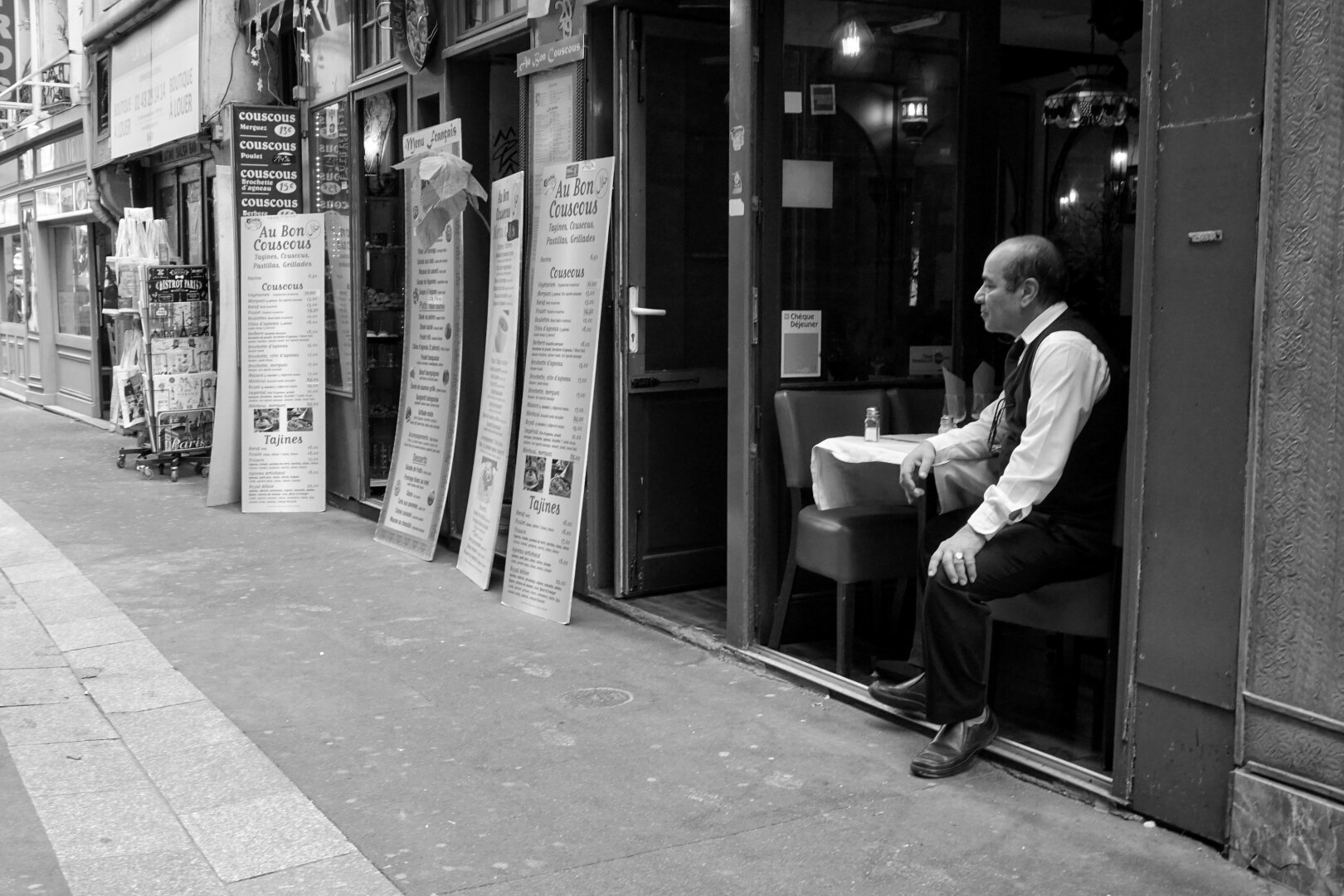 Black and white photo of a city street. In the foreground, a man dressed like a waiter sits at a cafe table, one foot hanging outside the cafe. Along the street are tall menus of other cafes and restaurants.