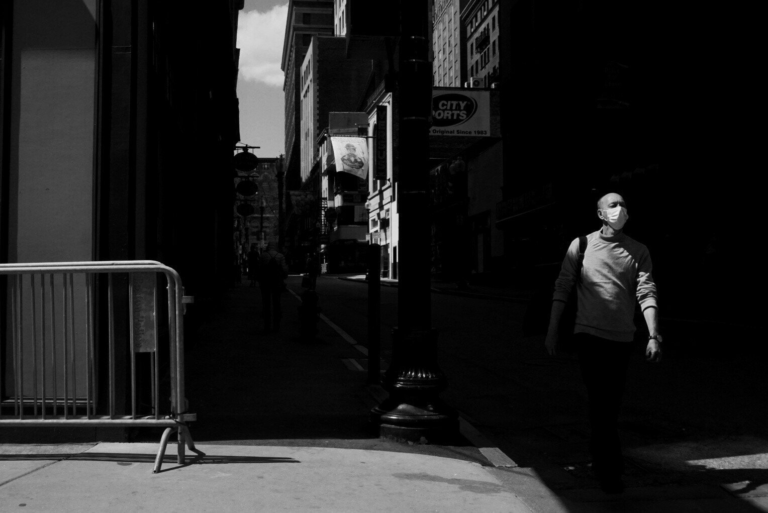 Black and white photo looking up a city street from the cross street. Most of the scene is hidden in shadow; tall buildings are sunlit in the background. To the right of the frame, a man in a surgical mask emerges from the shadows, his lower half still hidden.