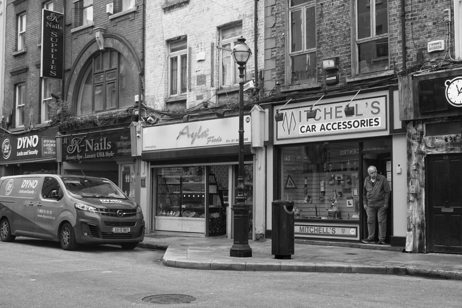 Black and white photo across a city street. A van is parked to the left of the frame, and the street is lined with shops and a nail salon. A man stands in the doorway of one of the shops.