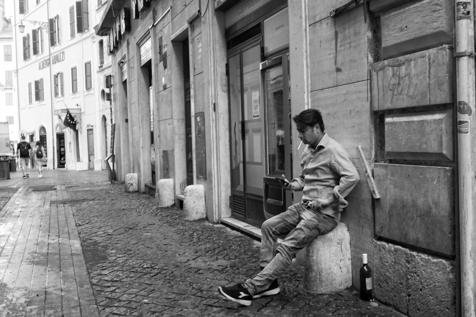 Black and white photo of a cobbled street. In the foreground, a young man sits on a concrete cylinder, one leg stretched out in front of him. He smoked a cigarette and looks at his phone. A bottle stands on the ground next to him.