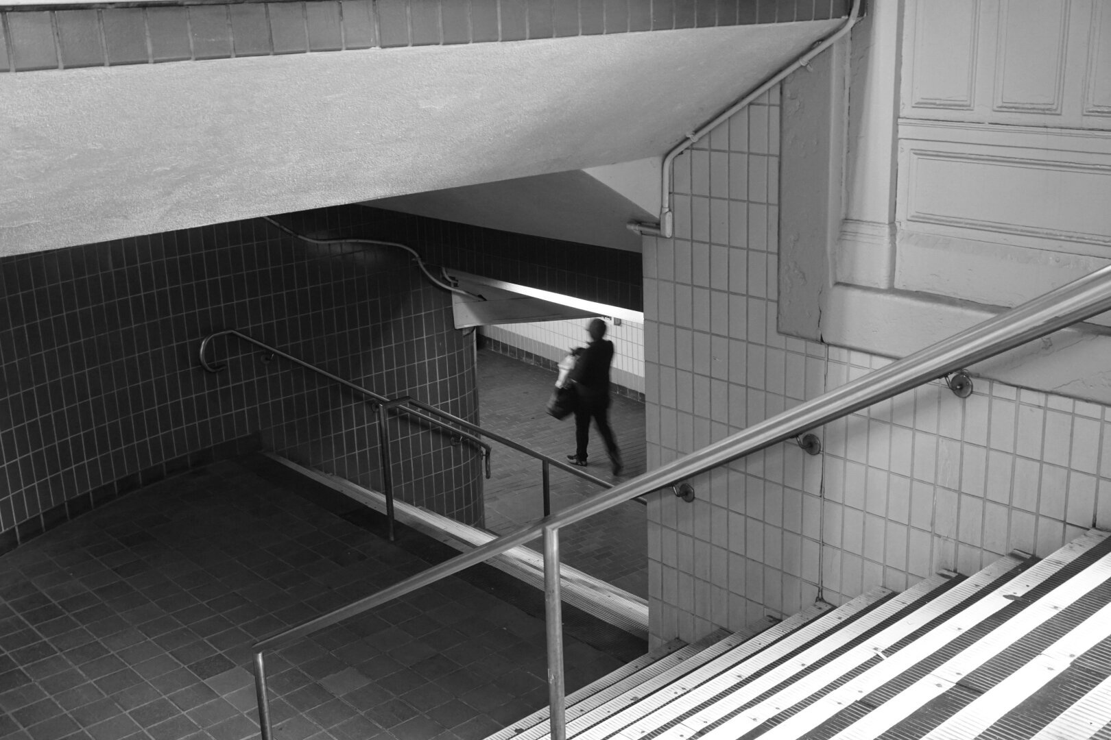 Black and white photo of the entrance to a subway station. There are two flights of stairs at a right angle to reach other. The banisters, stairs, walls, and ceiling form a pattern of diagonals that converges on a person walking past down below, a small figure in the pattern.