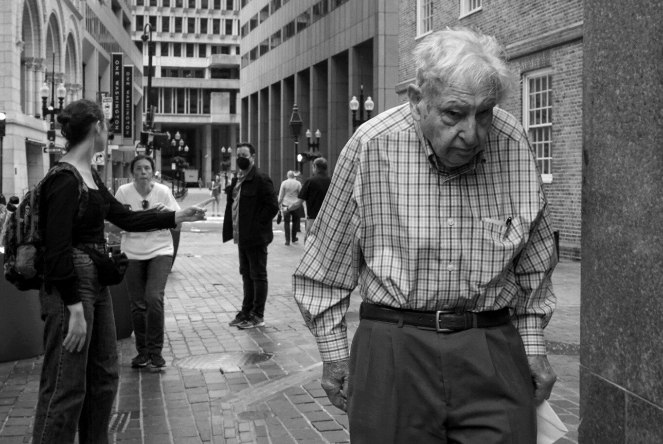 Black and white photo of a street scene. In the far background is Boston's brutalist city hall, and closer to us are people milling around on the brick sidewalk. Closest to us is an elderly man walking with his head down. He wears a plaid shirt and his pants are belted high on his waist.