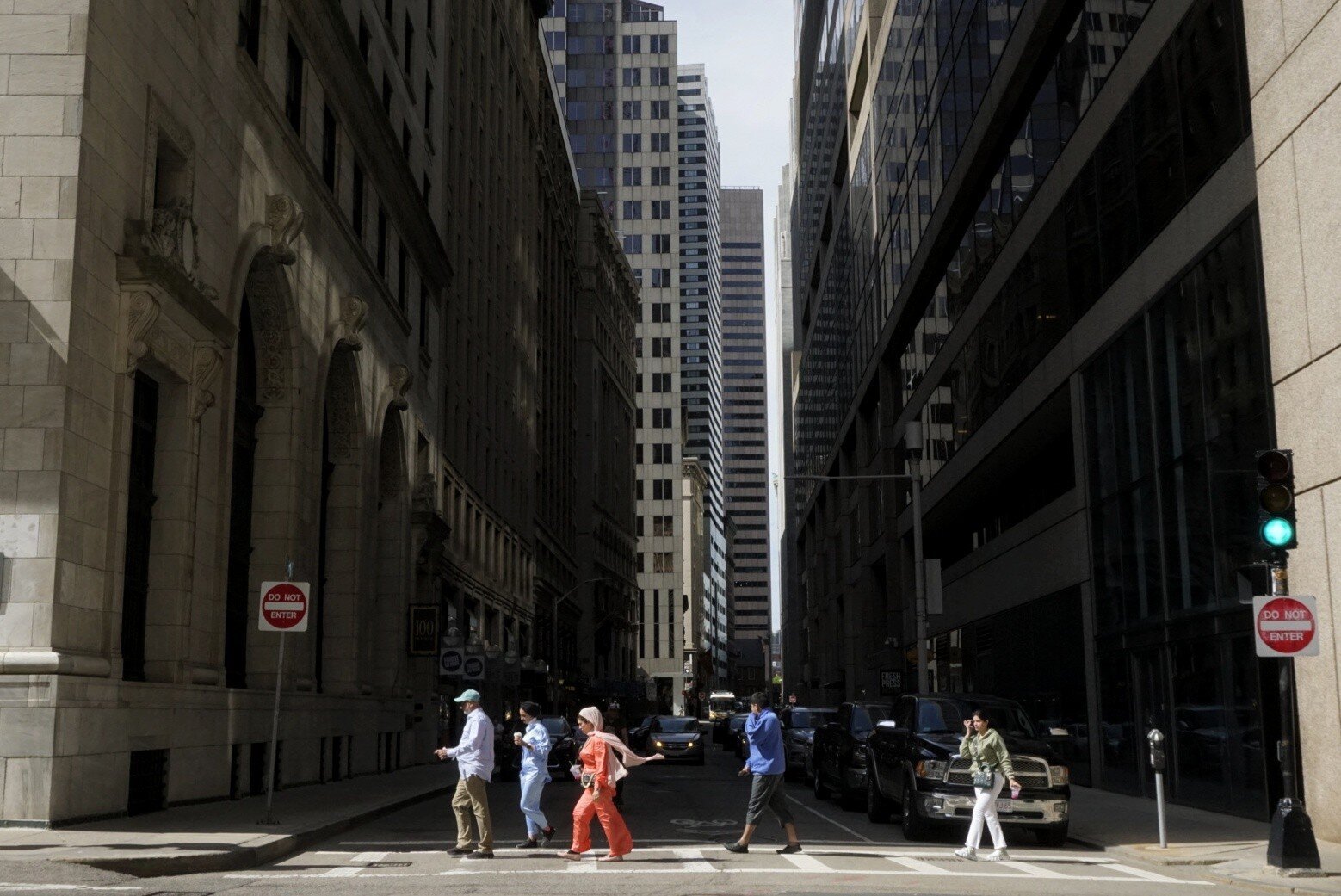 Color photo looking down a city street lined with tall buildings partly hidden in shadow. People walk single-file across a crosswalk, appearing small in the context of the buildings rising from all sides. They wear outfits of varying colors