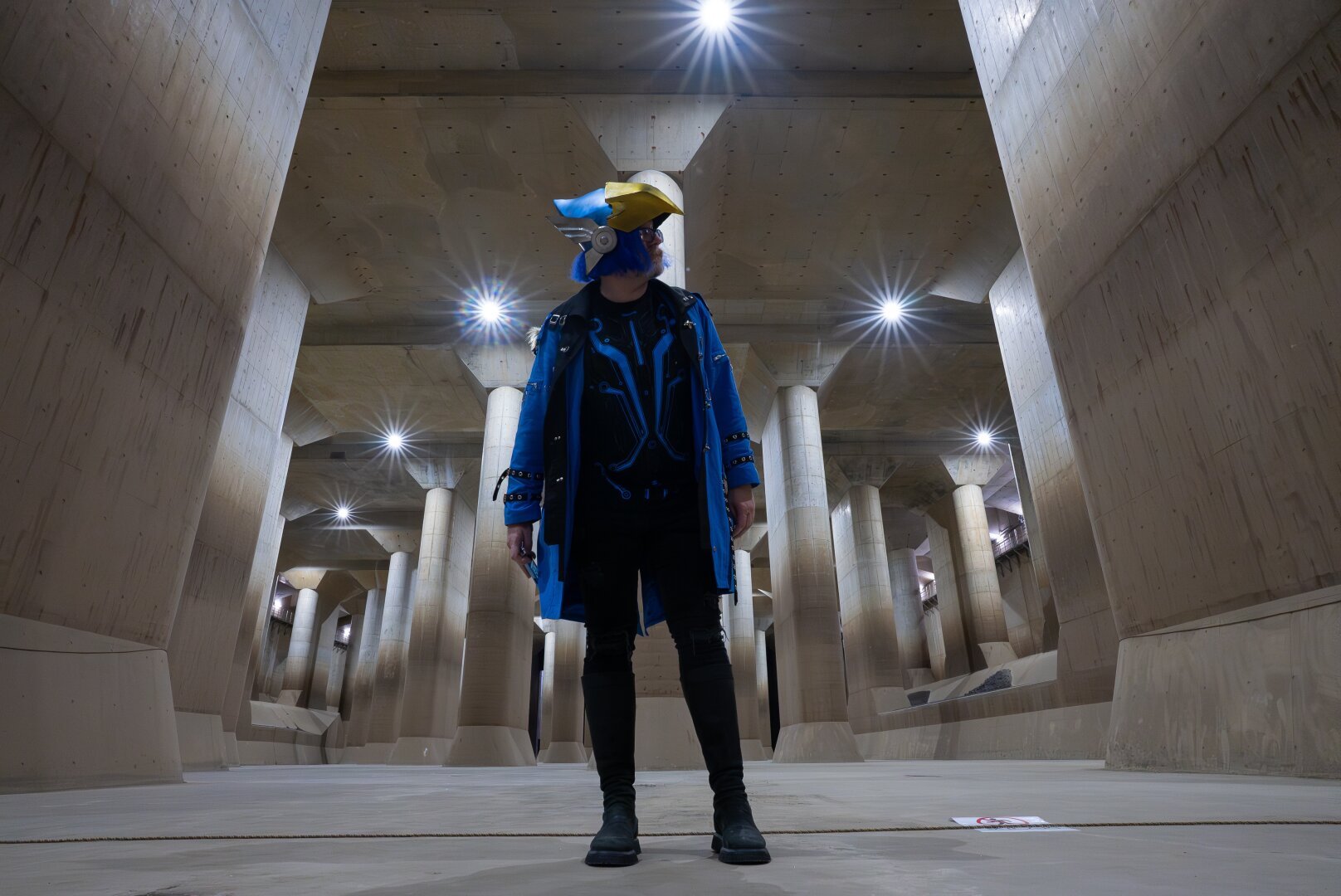 Person wearing a blue coat, a foam Pharah helmet, and black jeans, standing in the temple of the G-Cans project. The temple is 25 meters tall, 177 meters deep, and 78 meters wide. It has giant cement columns, and is lit by white lights on the ceiling.