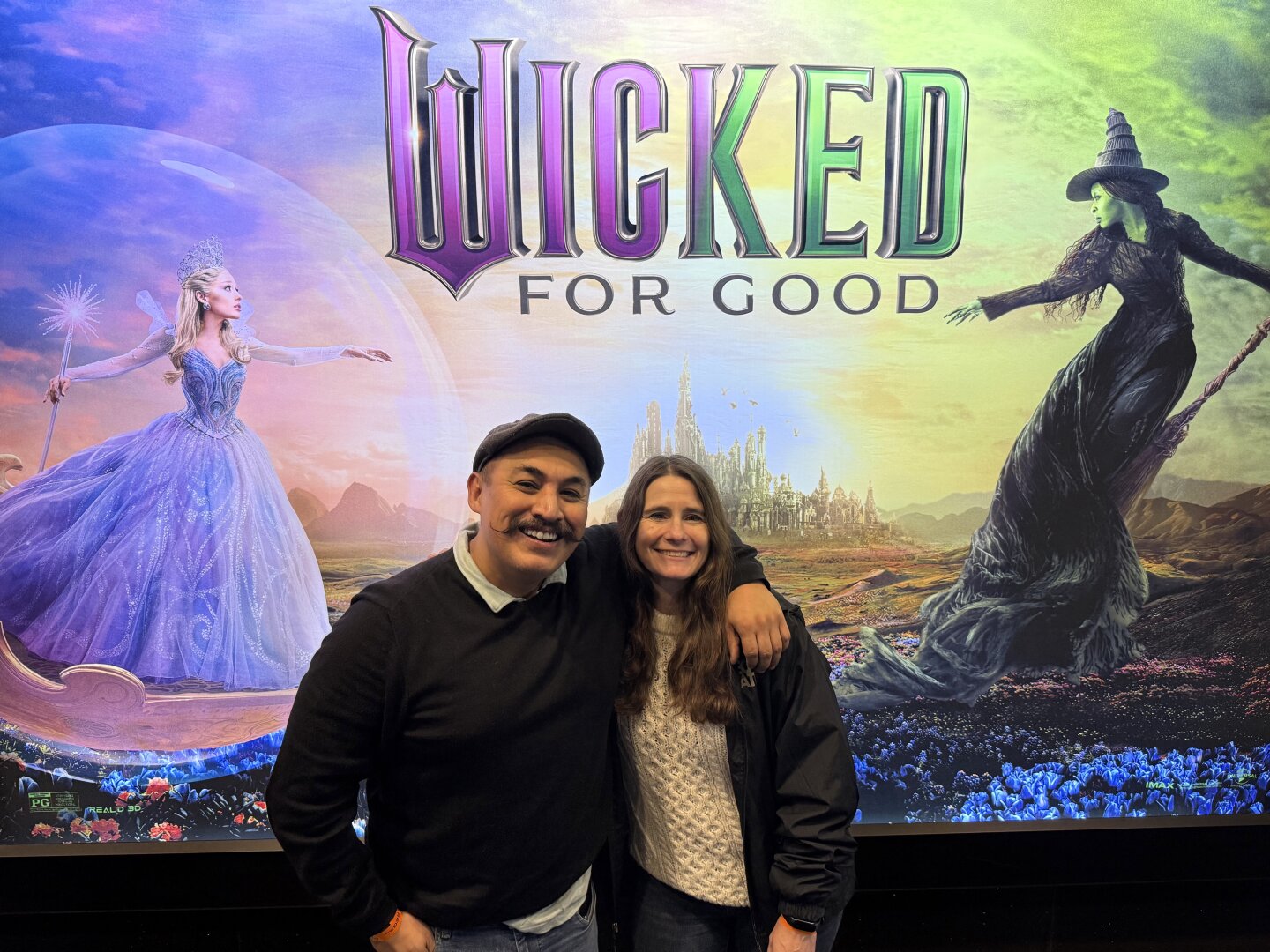 Two people in front of a Wicked poster