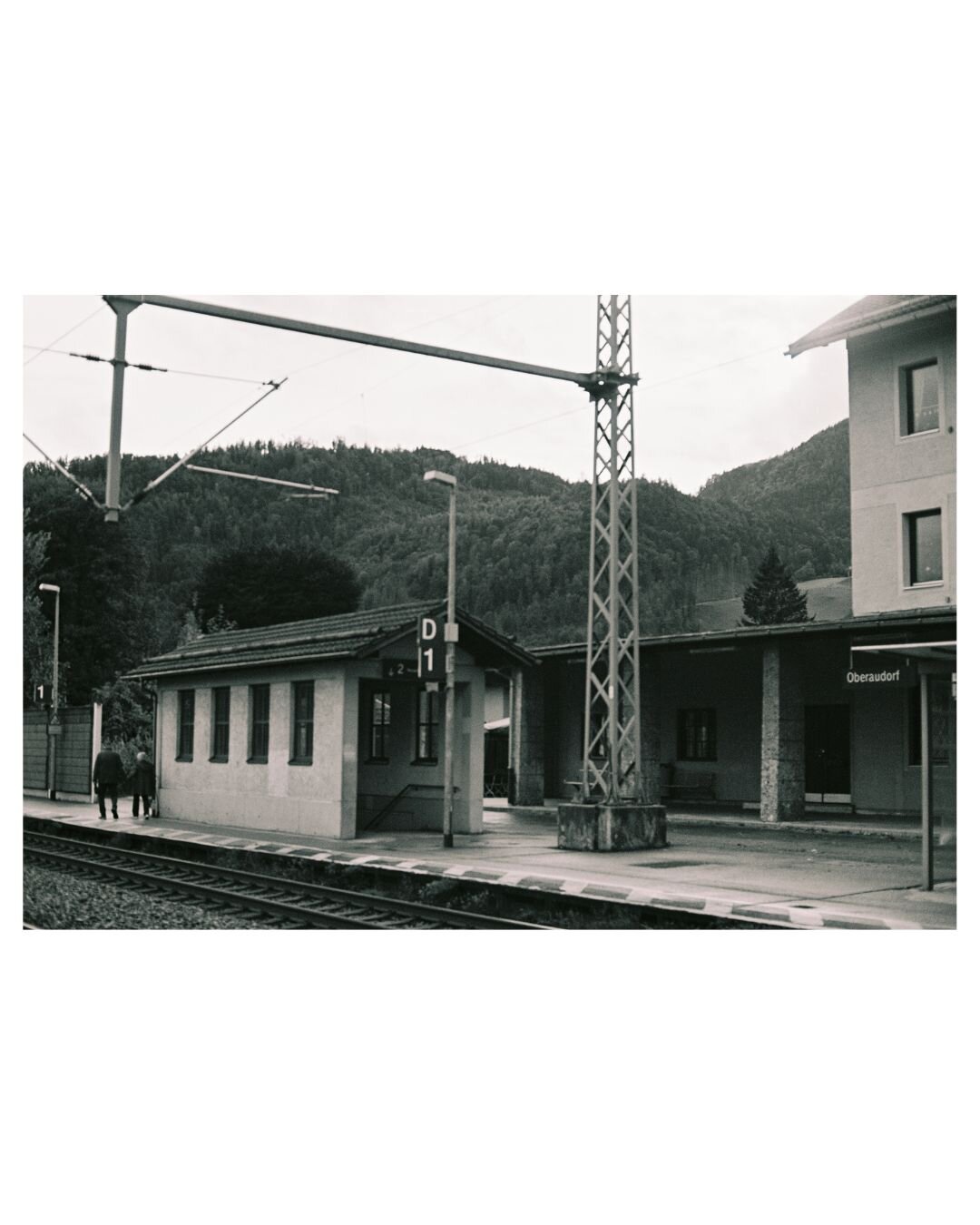 black and white photograph of a small village train station. An old couple is walking along platform D1 away from the camera; they are holding hands. Behind the train station you see wooded hills