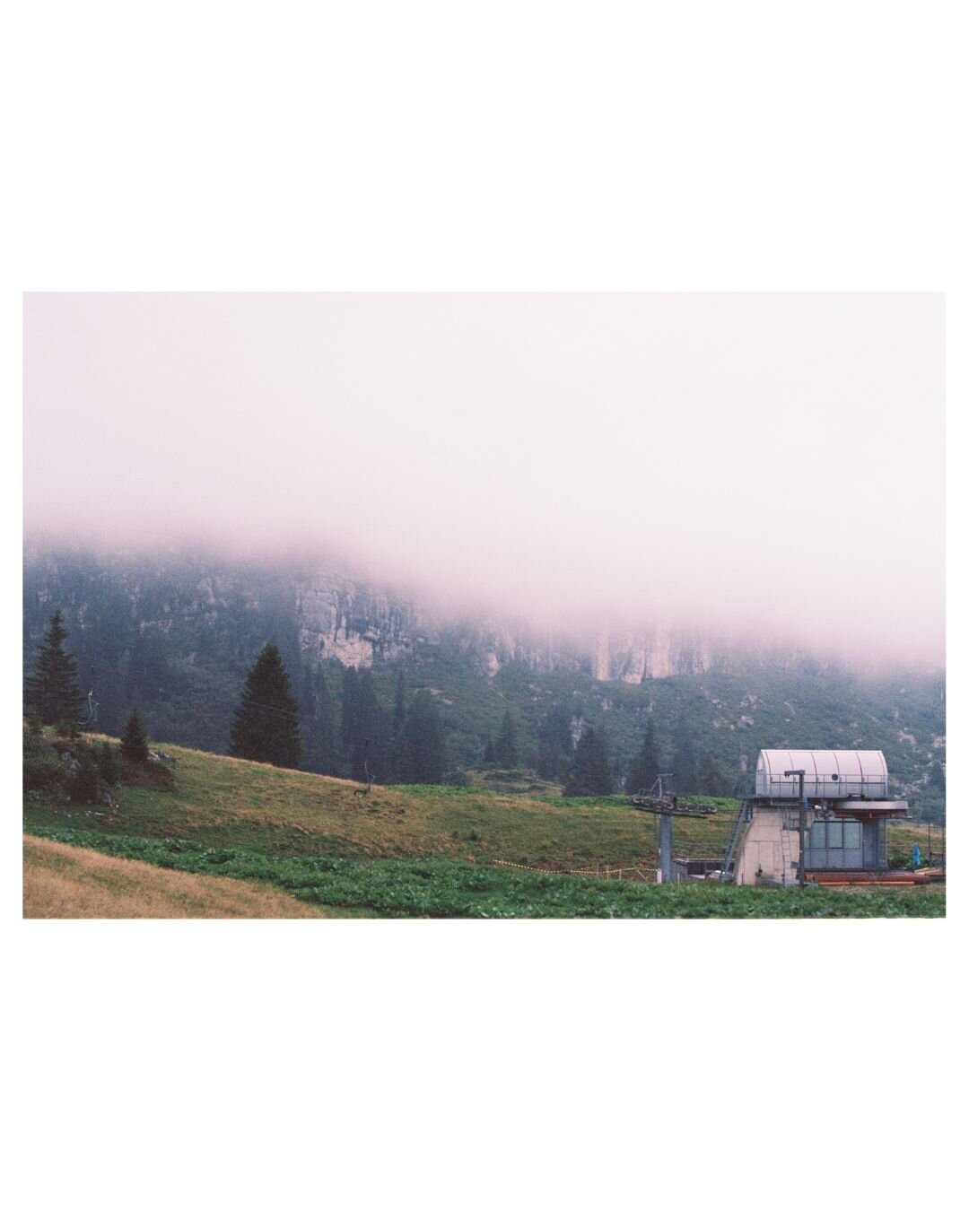 Green pastures, a small ski lift in the right corner of the picture. In the background the clouds are hanging deep and covering the rock walls almost completely
