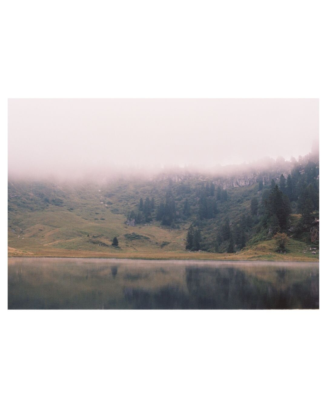 In the front a dark lake with the reflection of the background steep pastures with conifers. The mist is crawling over the rockwalls.