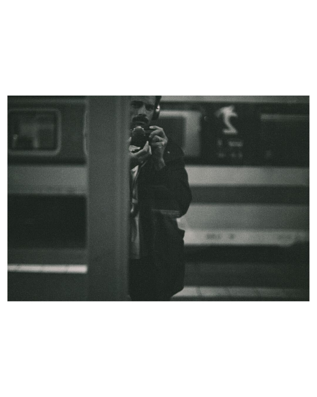 black and white photograph of myself in front of a mirror. very dark and grainy photo. on the left side you see a train on the background platform, in the middle I'm looking with my camera and headphones around a vertical metal construction that is cutting me in half, followed by the mirror reflection an other train on the right hand side of the photo.