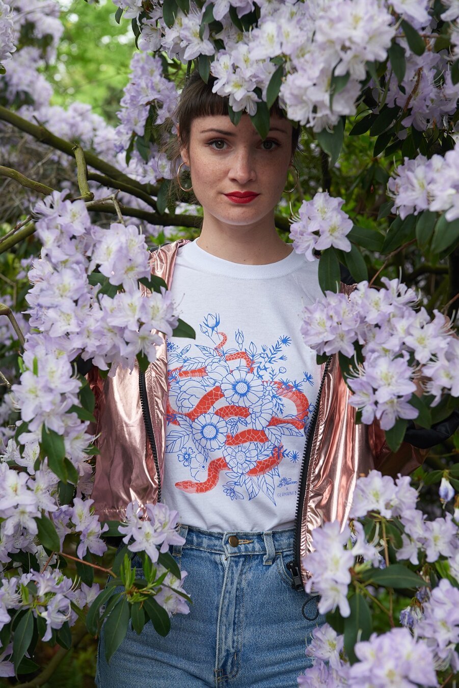 Photographie d'une femme cachée dans les branches fleuries d'un arbre. Elle porte un teeshirt avec un serpent et des fleurs. Le motif est sérigraphié en deux couleurs. Le serpent est orange et les fleurs bleu outre-mer.