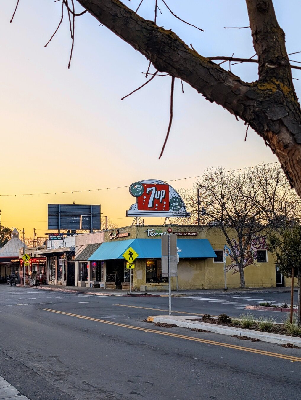 A photo of a vintage 7-Up sign in a small town at sunset. The sign is mounted on the roof of a building, and its bright colors are illuminated by the fading daylight. The building itself is a one-story brick structure with a flat roof and a row of windows. The street in front of the building is lined with trees and parked cars, and there are a few people walking by.