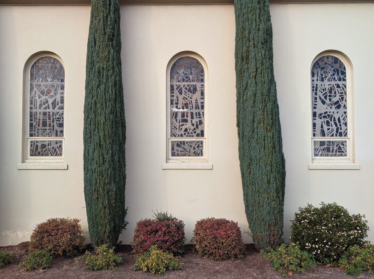 The image shows a building with three stained glass windows. The windows are arched and have a white frame. The stained glass is a blue-green color and has a variety of geometric shapes. There are two evergreen trees in front of the building, one on either side of the windows. There are also three small bushes in front of the building, one in front of each window. The ground is cOvered with dirt and there is some green grass in the lower left corner of the image.