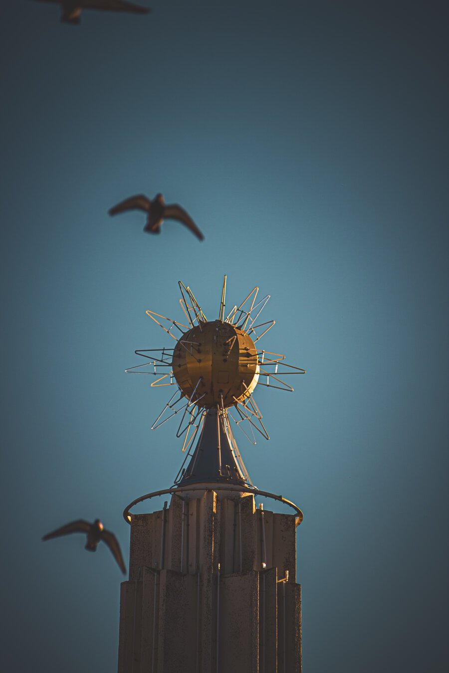 The image shows a close-up, slightly angled view of the top portion of a tall tower. The most prominent feature is a large, golden-colored sphere that resembles a stylized sun or starburst. It's made up of a central round sphere with numerous thin, radiating spikes or rods extending outwards, creating a star-like effect. Below the sphere is a slightly narrower, conical tower structure, textured with vertical lines or ribs. It appears to be made of panels or sections. The tower narrows as it goes down, leading the eye towards the bottom of the image. Surrounding the tower, and visible against a clear, light blue sky, are several birds in flight. They are silhouetted against the sky, appearing as dark shapes with outstretched wings. The birds are scattered, some above and some to the sides of the tower, adding a sense of movement to the image.