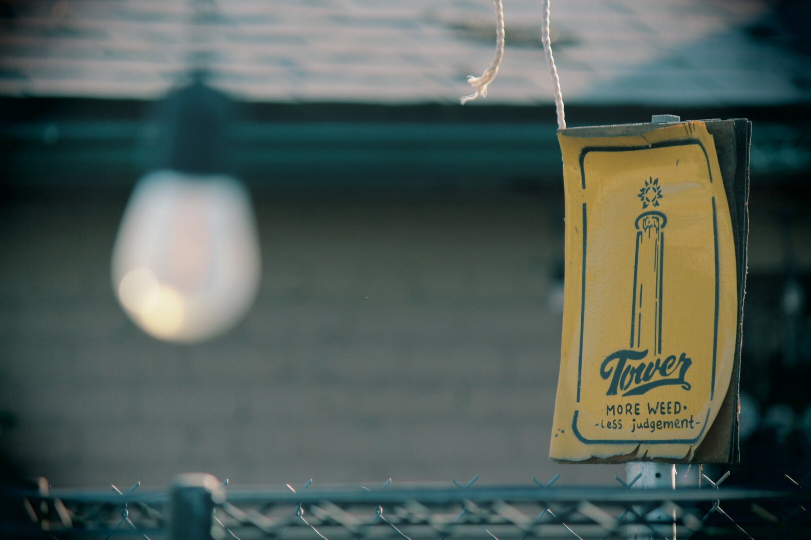 Outside, and it seems to be a bit overcast.  In the foreground, slightly to the right, is a sign. It's not a big, flashy sign, but rather a rectangular, kind of weathered one.  Made of thin metal, and it's a faded yellow color.
There's a small stylized illustration of the Tower Theater.
Below that symbol are two words stacked on top of each other. The word at the bottom is 