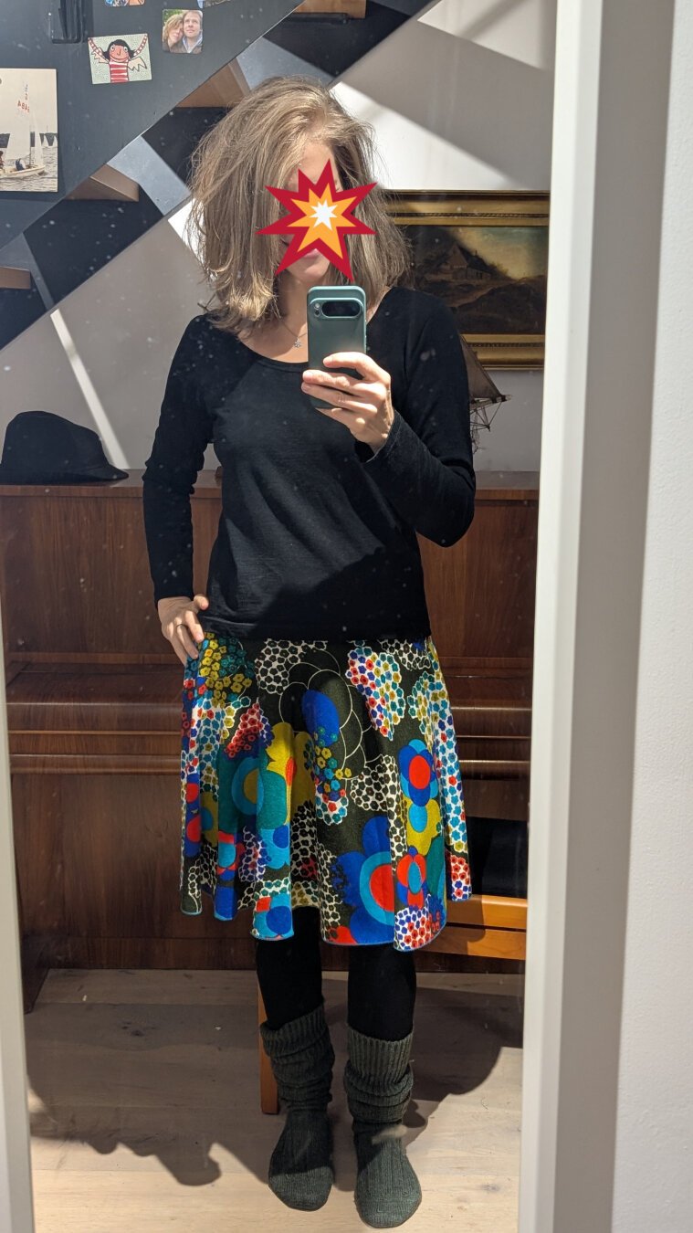 Spiegelsefie einer Frau mit schwarzem Pullover und bunten Tellerrock. Der Rock hat ein wildes Retro-Blumen-Muster in knalligen Farben Sie trägt eine schwarze Strumpfhose und grüne Wollstrümpfe.
Ihr Gesicht ist durch ein kleines Explosions-Emoji unkenntlich gemacht.