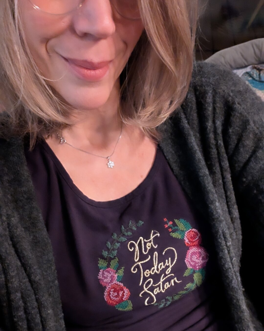 English below:
Selfie einer Frau mit grau-blondem Haar und Brille, die ein dunkel-lilanes Shirt trägt. Auf dem Shirt ist eine Stickerei mit bunten Blumen, in deren Mitte 