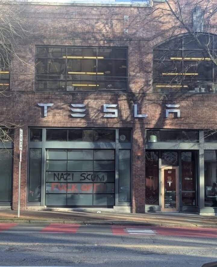 A Tesla storefront in an urban setting with a brick facade and large glass windows. The Tesla logo is prominently displayed above the entrance. On one of the lower garage-style windows, graffiti in bold black and red letters reads: 