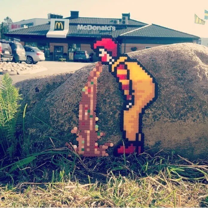 A pixelated Perler bead artwork by Swedish street artist Pappas Pärlor, depicting a glitched-out Ronald McDonald vomiting pixelated fast food onto a rock. The piece is placed outdoors in front of a McDonald's restaurant, blending humor and satire with urban surroundings. The artwork critiques fast food culture while embracing a nostalgic, video-game-inspired aesthetic.