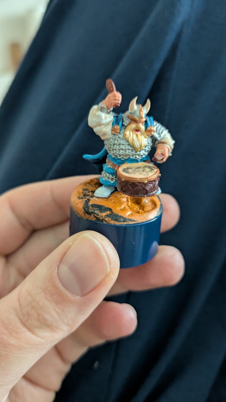 Ich halte im der Hand eine Miniatur eines Zwerges. Dieser hat ein Kettenhemd und einen gehörnten Helm auf dem Kopf. Er trägt einen Umhang und hat einen langen Bart. Er trommelt gerade auf einer Trommel, die vor seinem Bauch baumelt, wobei ein Arm mit Schlägel überm Kopf schwebt und der andere kurz vor der Trommel ist.