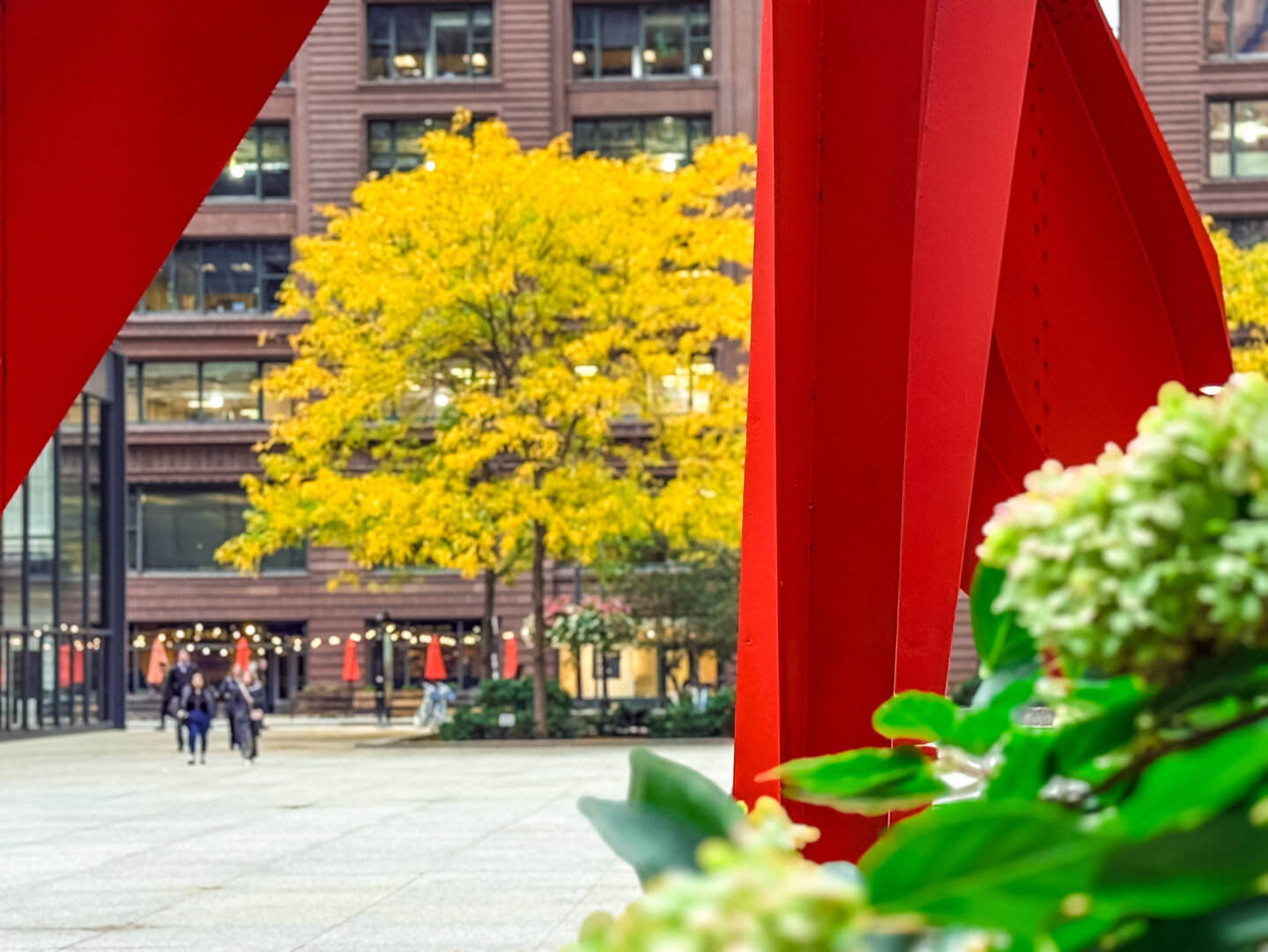 A vibrant red sculpture is prominently featured in the foreground, while in the background, a yellow-leaved tree and a plaza with people walking can be seen. The scene is framed by green foliage and decorative lights.