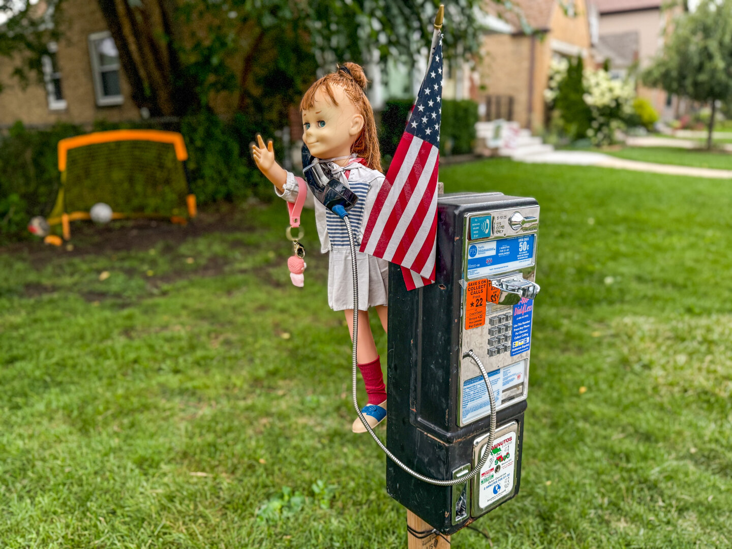A doll dressed in a striped dress and red socks is perched atop a payphone in a residential front yard. The doll holds a small American flag and a pink keychain, creating an eerie, whimsical scene. The payphone is covered in stickers and call instructions. In the background, there’s a grassy area with a soccer goal, a ball, and city houses under a cloudy sky.