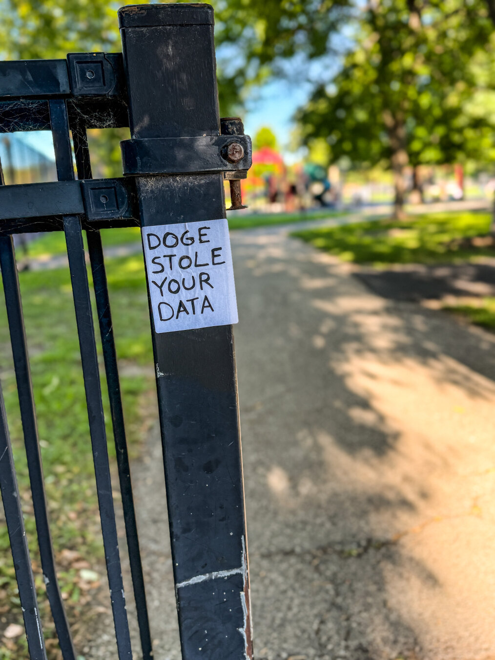 A black metal gate with a small handwritten sign that says "DOGE STOLE YOUR DATA." In the background, there is a pathway leading through a park with greenery and playground equipment visible.