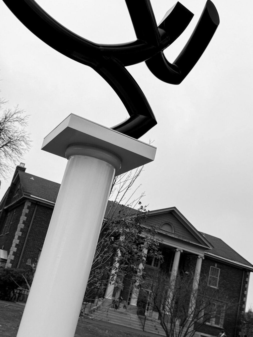 A close-up of a modern black sculpture set on a white pedestal, with a historic brick building in the background. The image is in black and white with a moody sky.