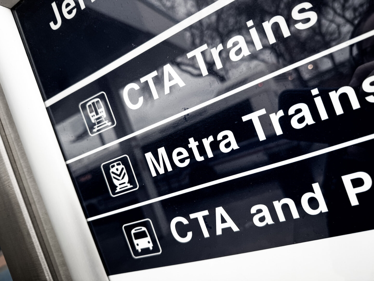 A close-up of a transit sign displaying information about CTA and Metra trains, along with corresponding icons. The sign is mounted on a white frame and shows a blurred background.