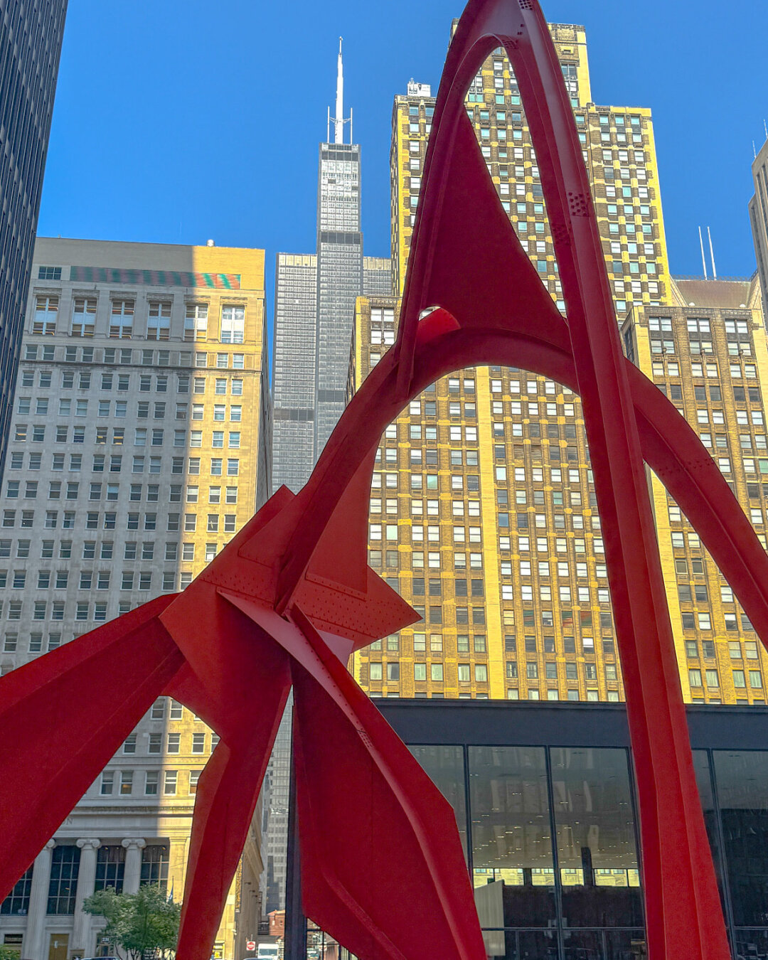 A large red sculpture stands in an urban plaza, flanked by tall buildings. The sculpture features dramatic, sweeping forms, contrasting with the surrounding architecture. A clear blue sky is visible overhead.