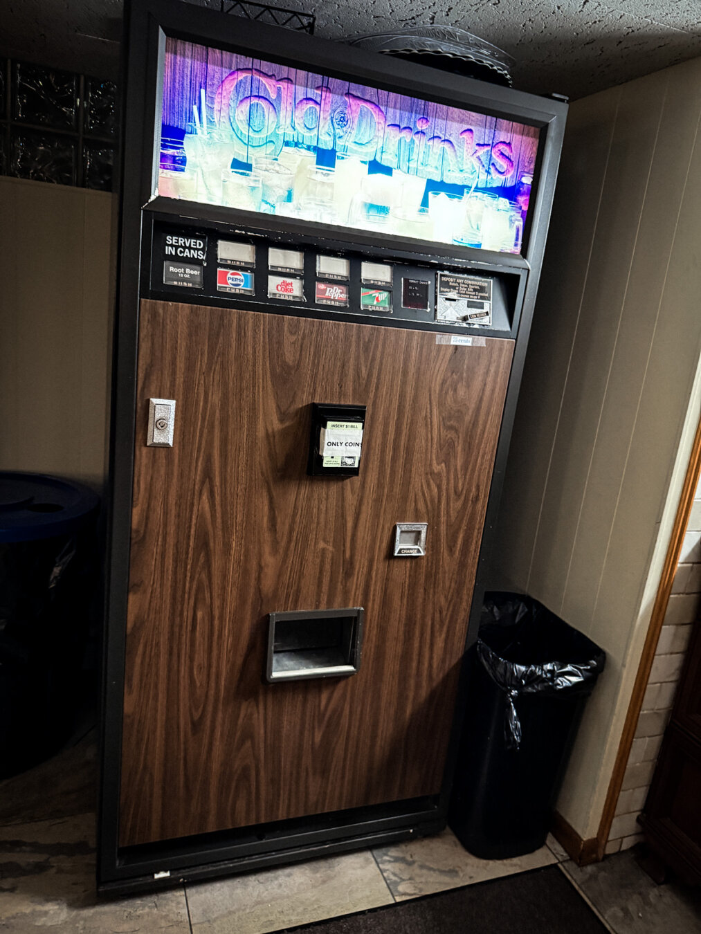 A vintage vending machine labeled "Cold Drinks," featuring various options for canned beverages like root beer and soda. The machine has a wooden design and illuminated signage, positioned in a corner next to a trash can.